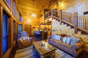 A log cabin interior with a wooden staircase, a living room with a sofa and coffee table, and a kitchen area. - Daydream Chalet · Hideaway: Nature Views, Kids Playroom & Cozy Balcony (Broken Bow)
