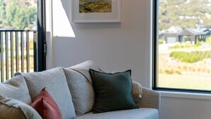 A beige sofa with multiple cushions, a framed landscape painting on the wall, and a view of a residential area through a large window. - Mountain View Apartment Steps from the Village (Wanaka)