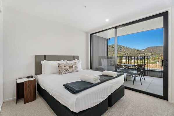 A modern hotel room with a large bed, a small side table, a balcony with a table and chairs, and a scenic view of hills. - Mountain View Apartment Steps from the Village (Wanaka)