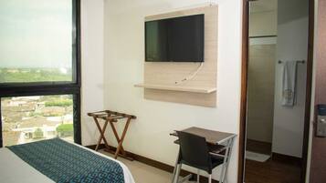 A hotel room with a bed, a desk, a chair, a TV mounted on the wall, and a view of the city through a large window.