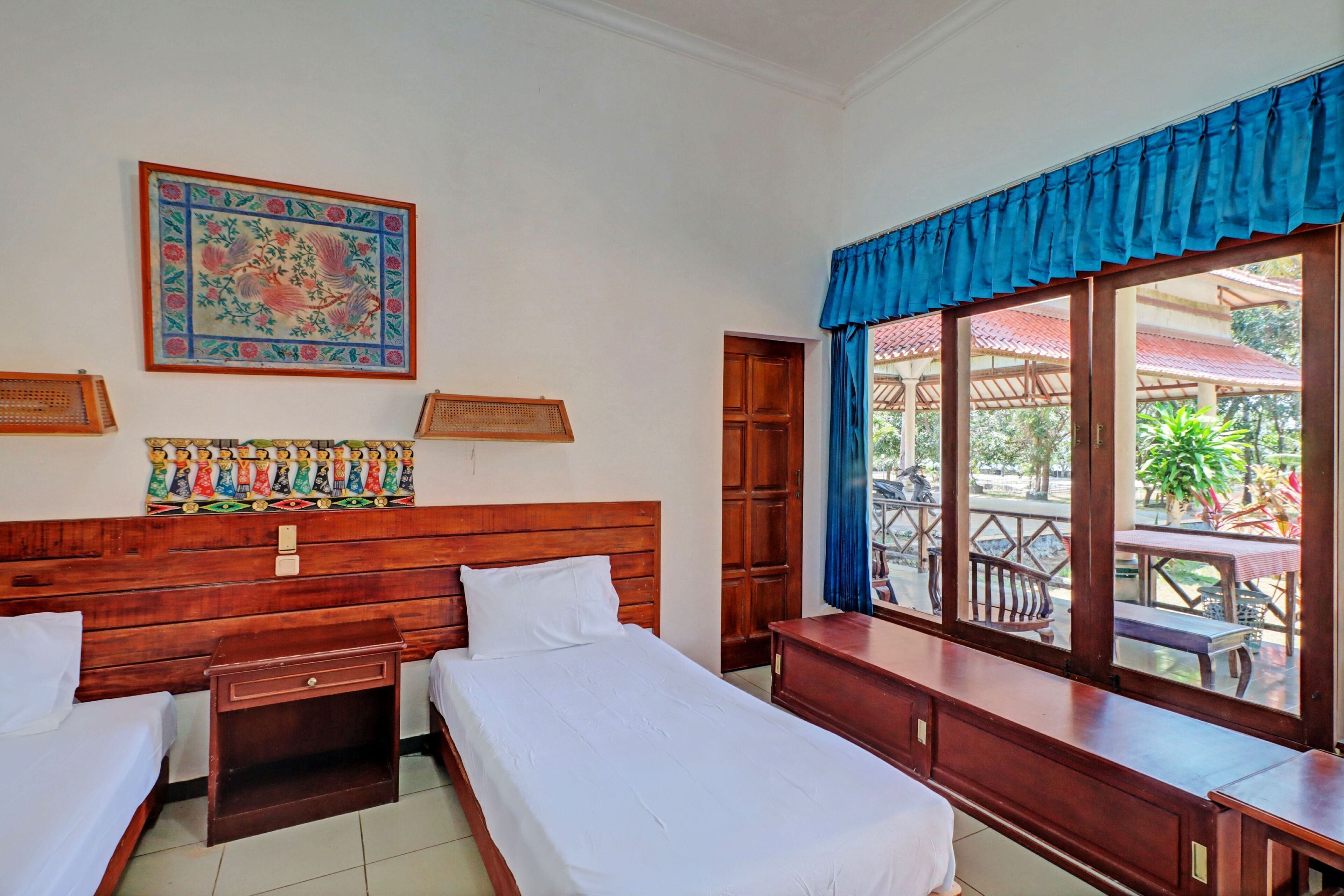 A hotel room with two beds, a wooden headboard, a large window with a blue valance, and a balcony with outdoor furniture.