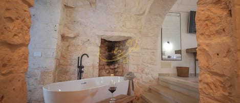 Un baño de paredes de piedra con una bañera independiente, un taburete de madera y una copa de vino.