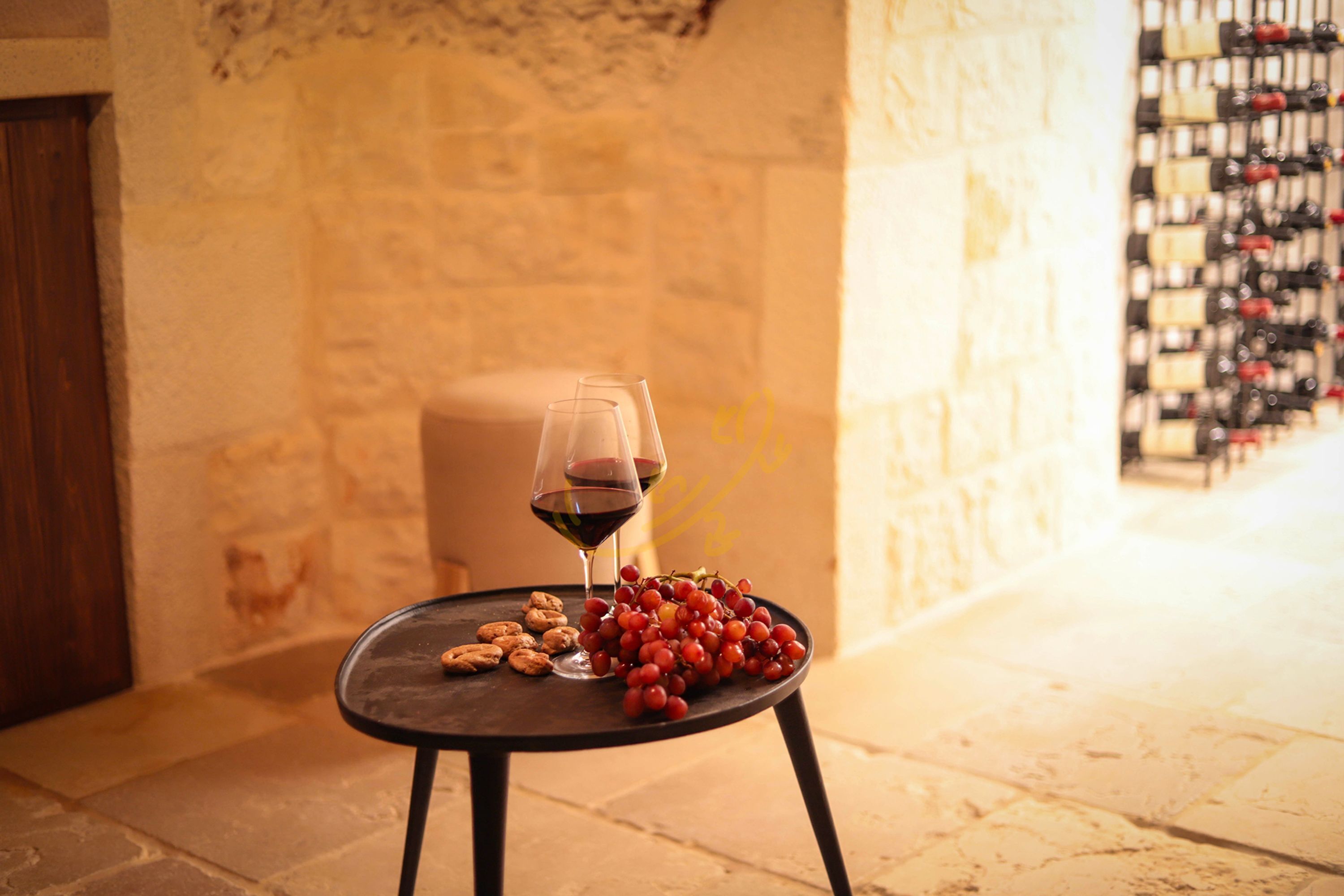 Una mesa con una copa de vino, uvas y galletas en una habitación de paredes de piedra con una vinoteca al fondo.