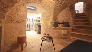 Um quarto com paredes de pedra, uma escada, uma pequena mesa com copos e uvas, e uma cadeira.