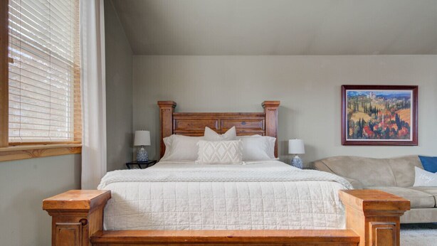 Un dormitorio con cama de madera, ropa de cama blanca, un sofá color beige y un cuadro en la pared.