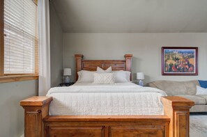Une chambre à coucher avec un lit en bois, du linge de lit blanc, un canapé beige et un tableau accroché au mur.