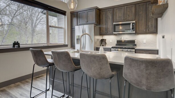 Une cuisine moderne avec des meubles en bois foncé, des appareils électroménagers en acier inoxydable et une grande fenêtre donnant sur une rue bordée d’arbres.
