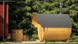 A wooden barrel-shaped structure with a chimney, a red building, and a wooden bench in a garden setting.