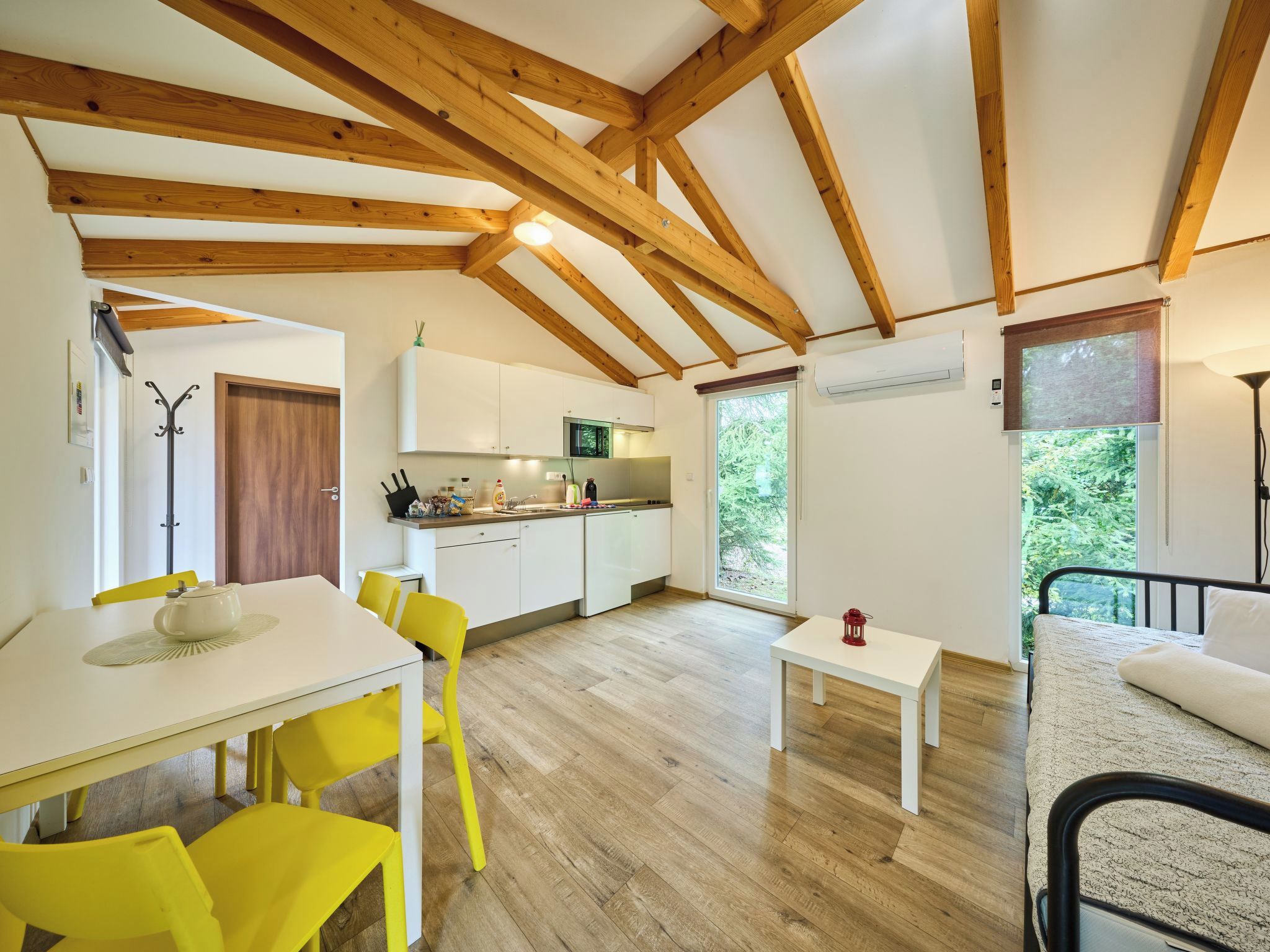 A modern kitchen with white cabinets, a microwave, and a sink. There is a dining area with a white table and yellow chairs. The room has wooden beams on the ceiling and large windows.