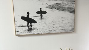 A framed photograph of surfers on a beach, a wooden dining table with black placemats, and potted plants on the table. - Norfolk Hideaway - Port Macquarie | Easy walk to Ocean and Beaches  (Port Macquarie)