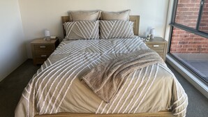 A bed with a striped bedspread and two pillows, a wooden nightstand with a lamp, and a window with a brick wall view. - Norfolk Hideaway - Port Macquarie | Easy walk to Ocean and Beaches  (Port Macquarie)