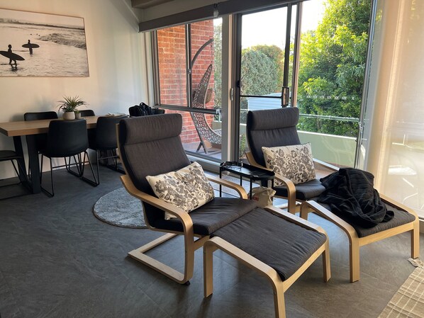 A modern living room with two lounge chairs, a dining table, and a large window offering a view of a brick building and greenery.
