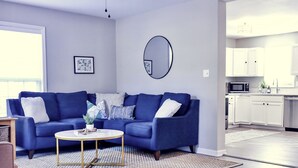 Un salon moderne avec un canapé bleu, une table basse ronde et un coin cuisine doté de meubles blancs et d’appareils électroménagers.