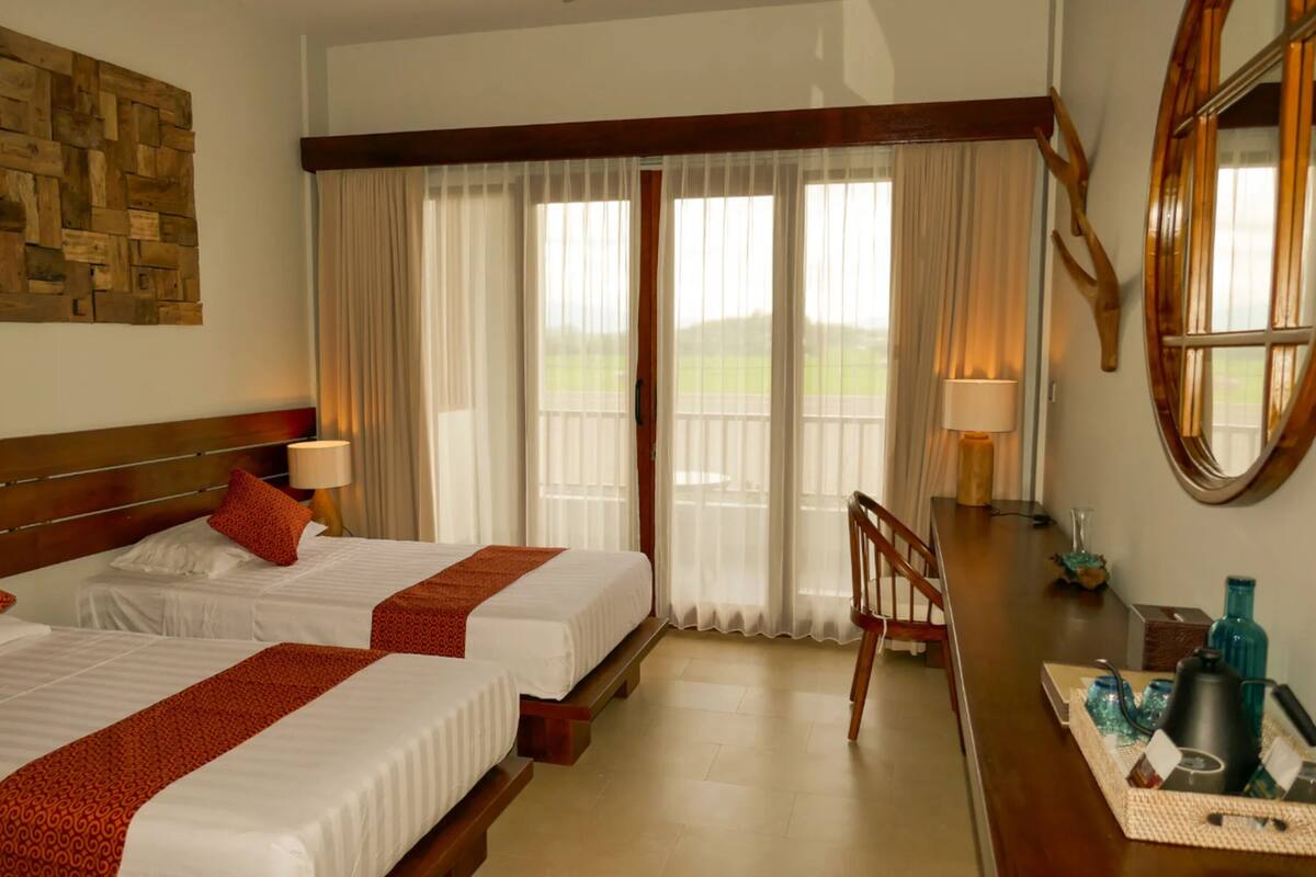 A hotel room with two beds, a wooden headboard, a balcony with curtains, a round mirror, and a small table with a lamp and decorative items.