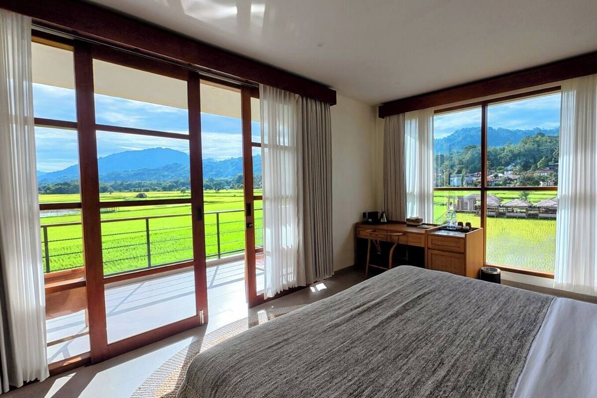 A hotel room with a large bed, a desk, and a view of a green landscape through large windows.