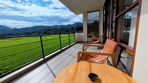 A balcony with a view of green fields and mountains, a wooden table, and two chairs. - Santai Toraja (Rantepao)