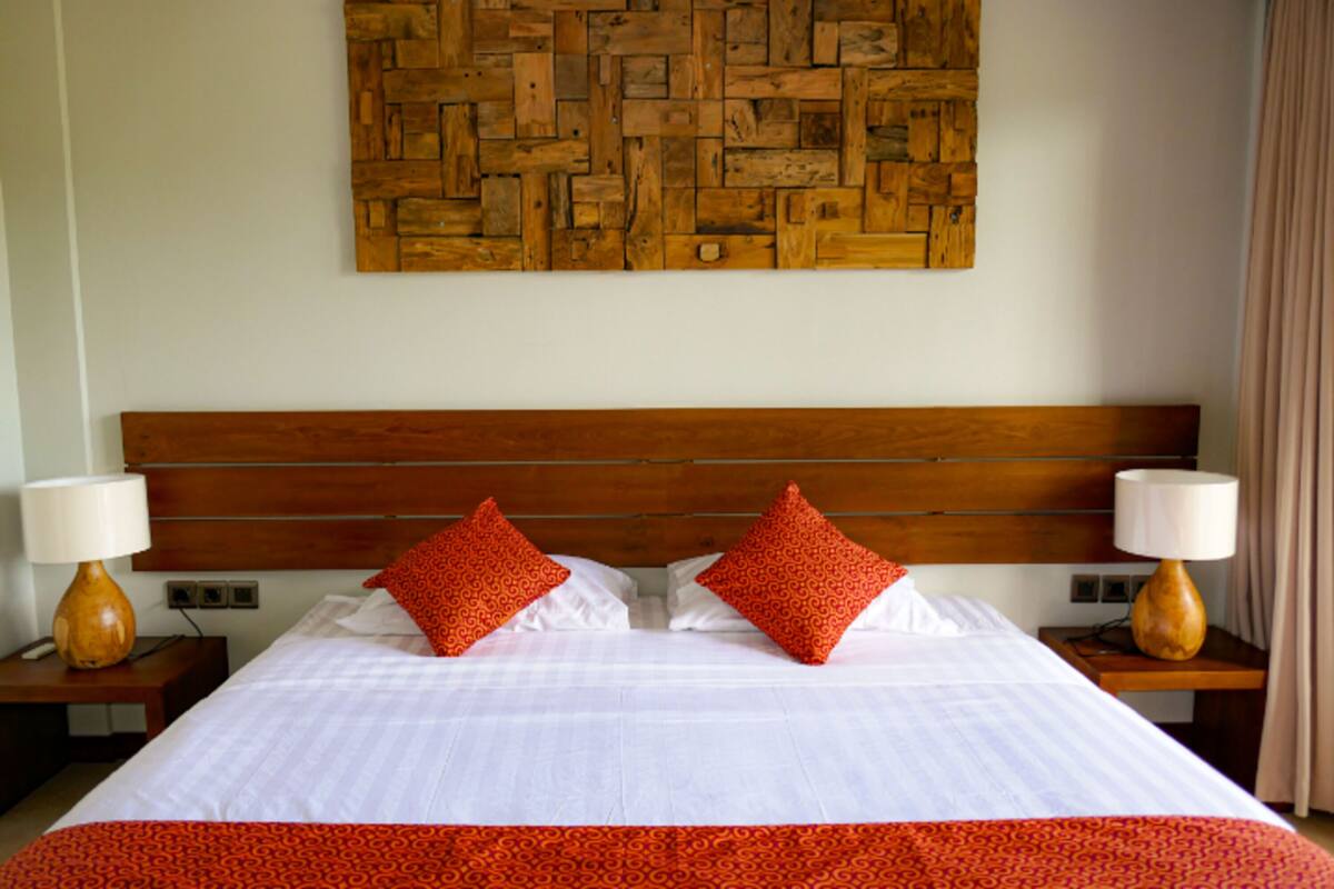 A hotel room with a wooden headboard, two red patterned pillows, white bedding, and two bedside lamps.