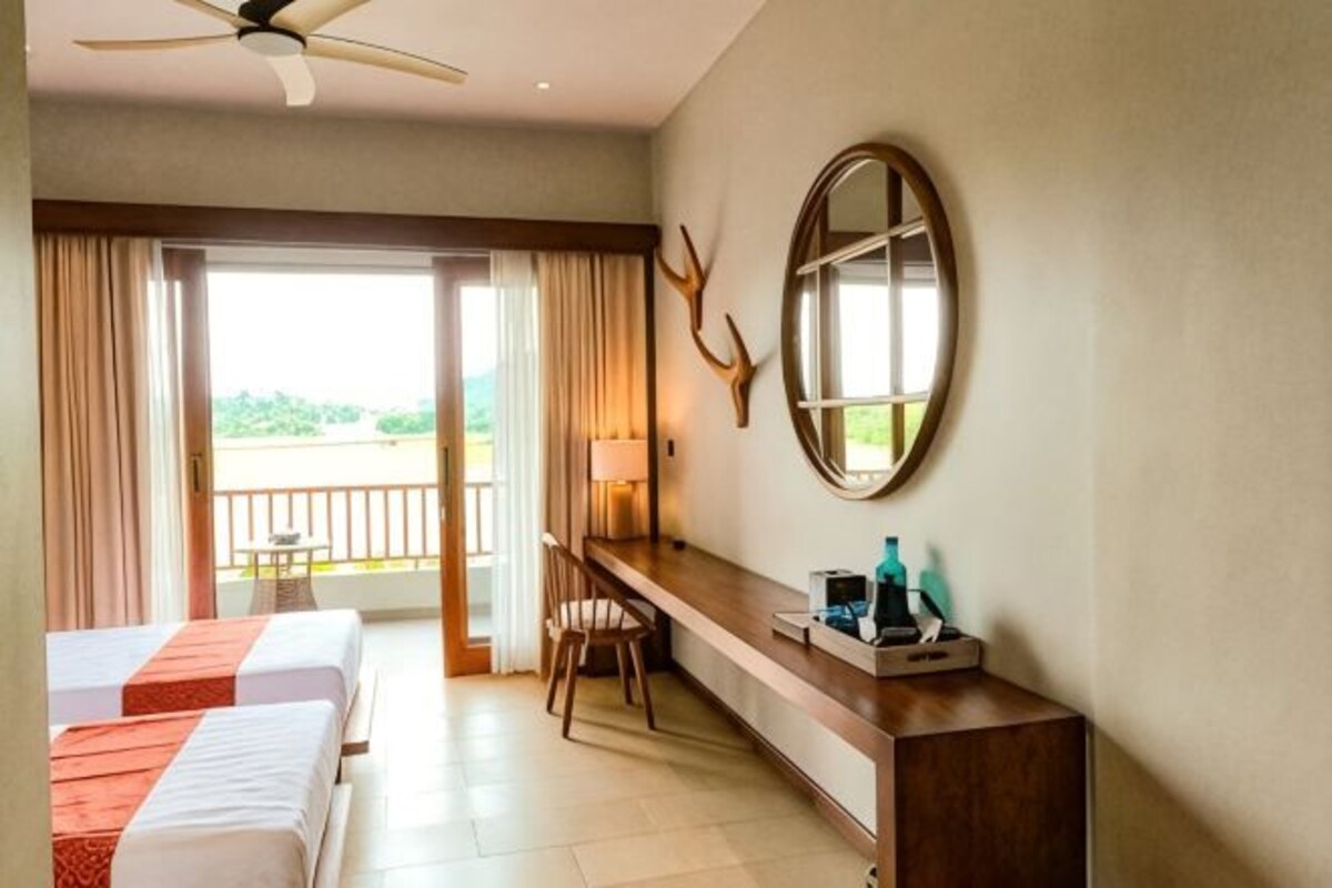 A hotel room with two beds, a desk, a chair, a mirror, and a balcony with curtains.