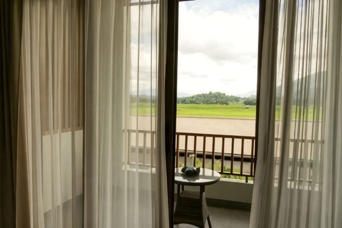A room with a view of a green field and a road, a small table with a chair, and sheer curtains.