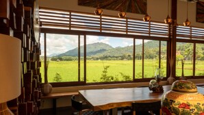 A room with a large window offering a view of a green field and mountains. There is a wooden table with chairs and a decorative vase. - Santai Toraja (Rantepao)