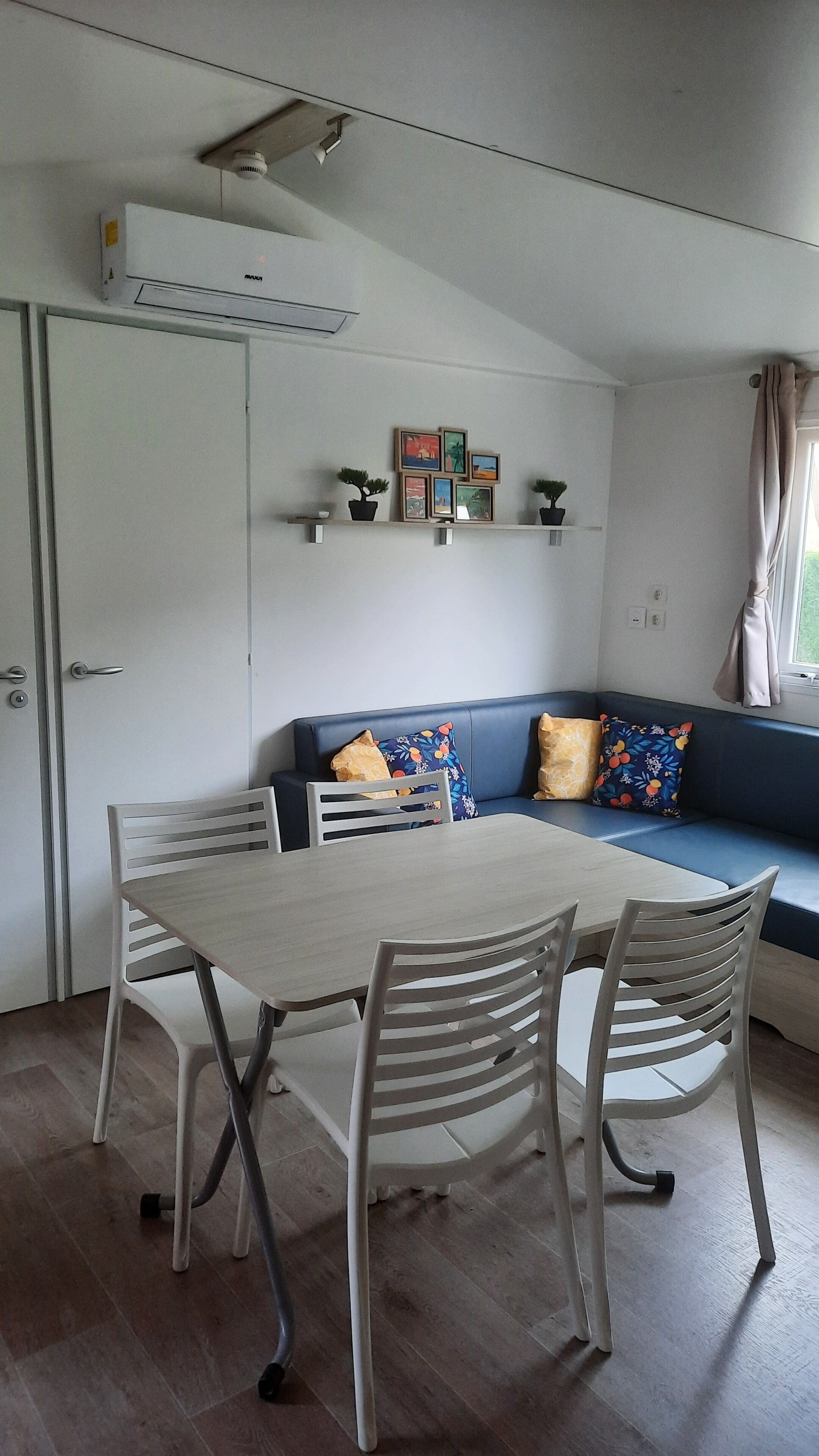 A compact living space with a dining table and chairs, a sofa with cushions, a wall-mounted shelf with framed pictures, and a window with curtains.