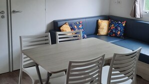 A compact living space with a dining table and chairs, a sofa with cushions, a wall-mounted shelf with framed pictures, and a window with curtains. - Joli Mobilhome Climatisé, 4 Pers. Campingla Falaise Plage à 300m, Piscine (Narbonne)