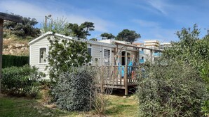 A small, single-story house with a deck and a slide, surrounded by dense shrubbery and trees. - Joli Mobilhome Climatisé, 4 Pers. Campingla Falaise Plage à 300m, Piscine (Narbonne)