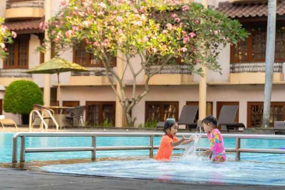 Un espace piscine où deux enfants jouent dans l’eau, entourés d’un bâtiment doté de balcons et d’un arbre aux fleurs roses.