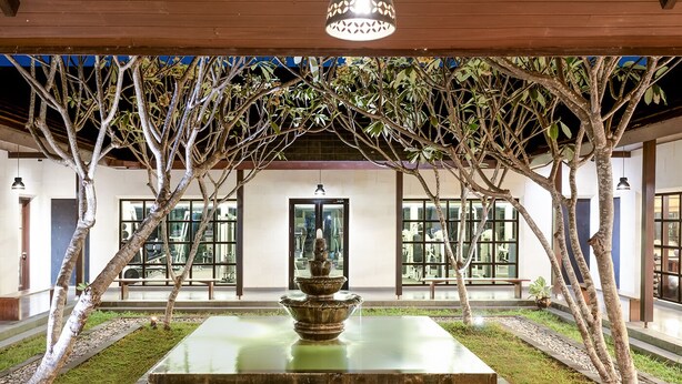 Une cour intérieure agrémentée d’une fontaine centrale, entourée d’arbres et bordée d’un bâtiment doté de grandes fenêtres.