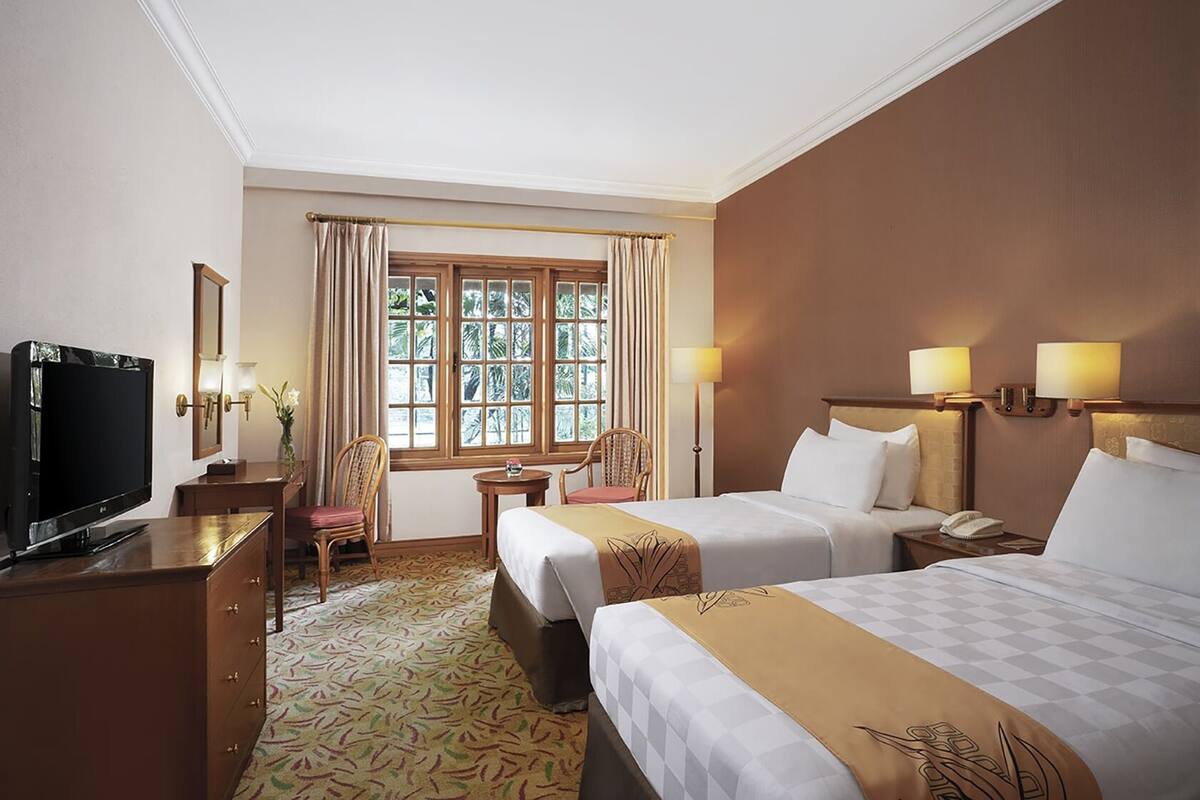 Habitación de hotel con dos camas, un escritorio con televisor, una silla, una mesita y una ventana con cortinas.