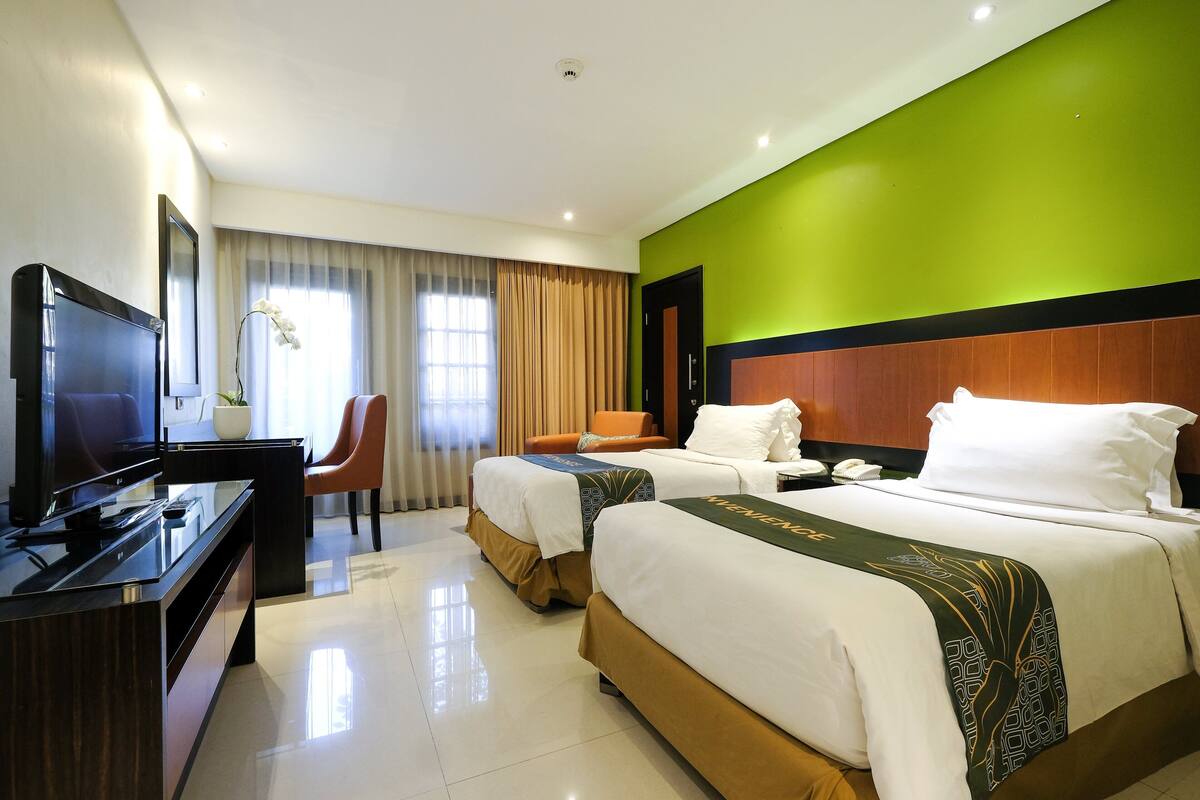 Habitación de hotel con dos camas, un escritorio con televisor, una silla y un ventanal con cortinas.