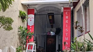 Entrada de un edificio con un cartel rojo que dice "MERRY CHRISTMAS", un taburete blanco pequeño y plantas en macetas.