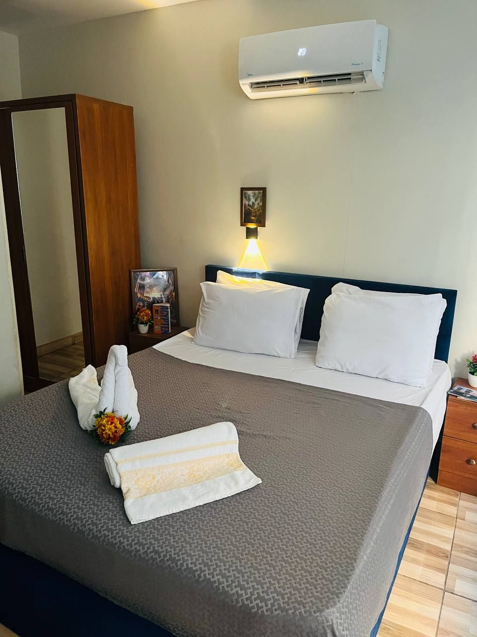 Un letto rifatto con lenzuola bianche, un condizionatore fissato al muro, una lampada da comodino e un quadro incorniciato appeso.