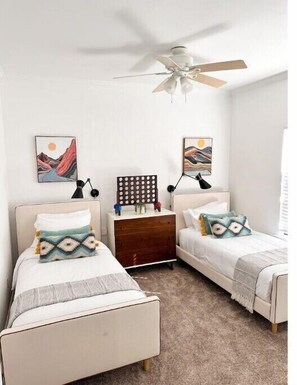 Un dormitorio con dos camas individuales, una mesita de noche de madera, un ventilador de techo y decoración en la pared.