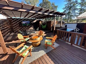 Una terraza de madera con mobiliario exterior, incluyendo sillas y una mesa redonda, bajo una pérgola.