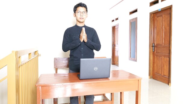 Uma pessoa em pé, perto de uma mesa de madeira, com um laptop, num espaço com portas de madeira e um radiador.