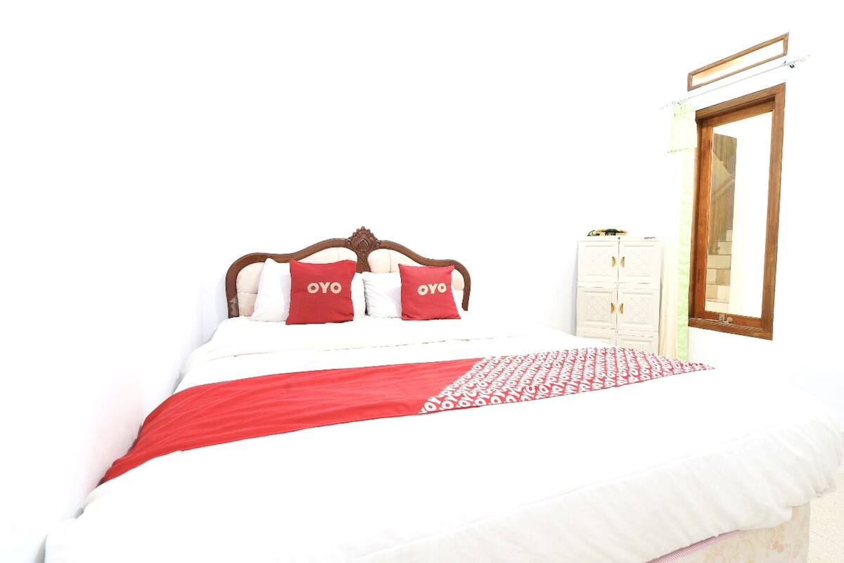 A bed with a white and red bedspread, two pillows with "OYO" text, a wooden headboard, a white wardrobe, and a mirror on the wall.