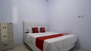 Cama bem arrumada com cabeceira branca e detalhes vermelhos, guarda-roupa branco com recortes circulares e uma parede branca simples com duas tomadas.
