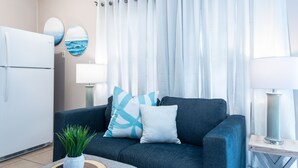 Un salon moderne avec un canapé bleu foncé, une petite table ronde ornée d’une plante, une lampe et un réfrigérateur blanc.