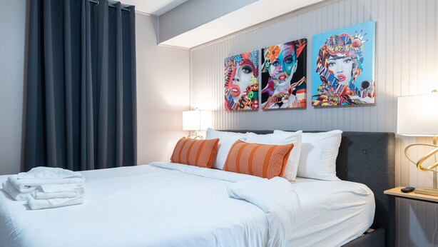 Un lit bien fait, avec du linge de lit blanc et des coussins orange, une tête de lit grise et deux tableaux accrochés au mur.