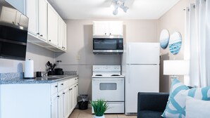 Une cuisine compacte avec des meubles blancs, un four à micro-ondes, une cuisinière et un réfrigérateur. On y trouve une petite plante posée sur un plateau en bois, un canapé gris avec des coussins bleus et une lampe fixée au mur.