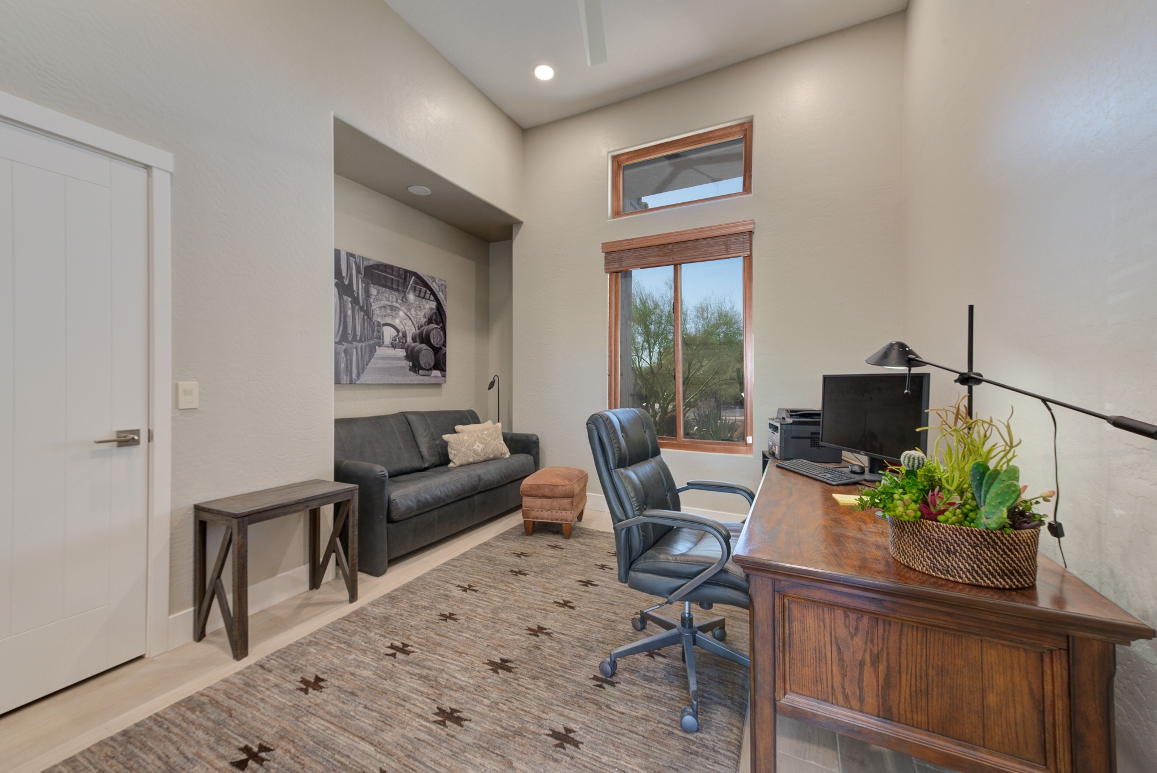 A modern living room with a black leather sofa, a wooden coffee table, a black office chair, a desk with a computer and a lamp, and a large window with a view of trees.