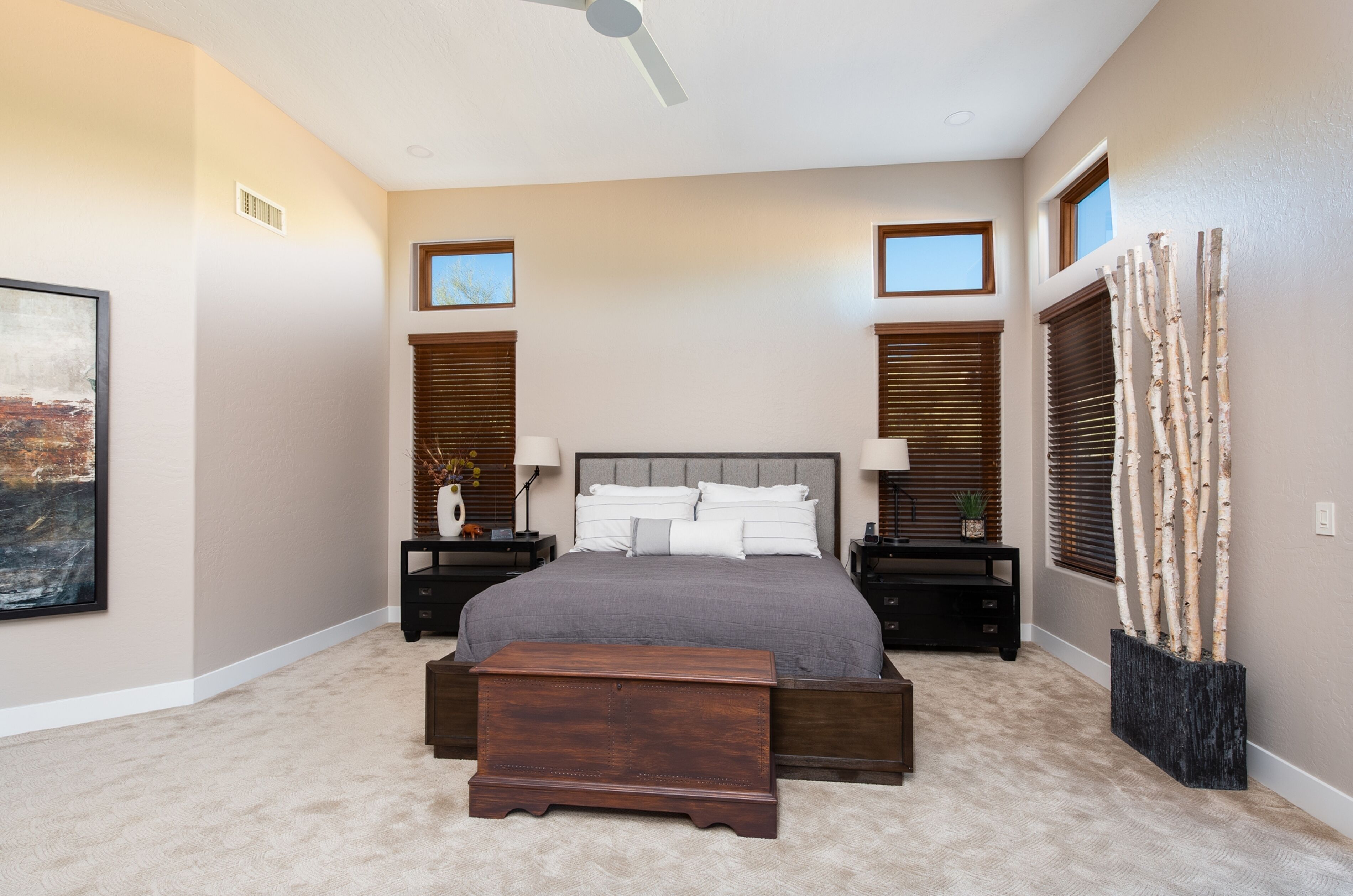 A bedroom with a large bed, two nightstands, a wooden bench, a ceiling fan, and a decorative tree.