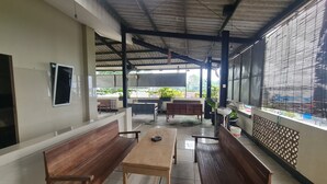 A covered outdoor seating area with wooden benches and tables, a tiled floor, and a view of the surrounding area.