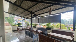A covered outdoor seating area with benches and tables, potted plants, and a view of the surrounding area.