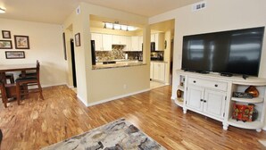 A spacious living room with hardwood flooring, a dining area with a wooden table and chairs, a kitchen with white cabinets, and a large flat-screen TV on a white cabinet. - SE Boise Condo • Pool • Parking • Hot Tub+ 2 Private Master Suites (Boise)