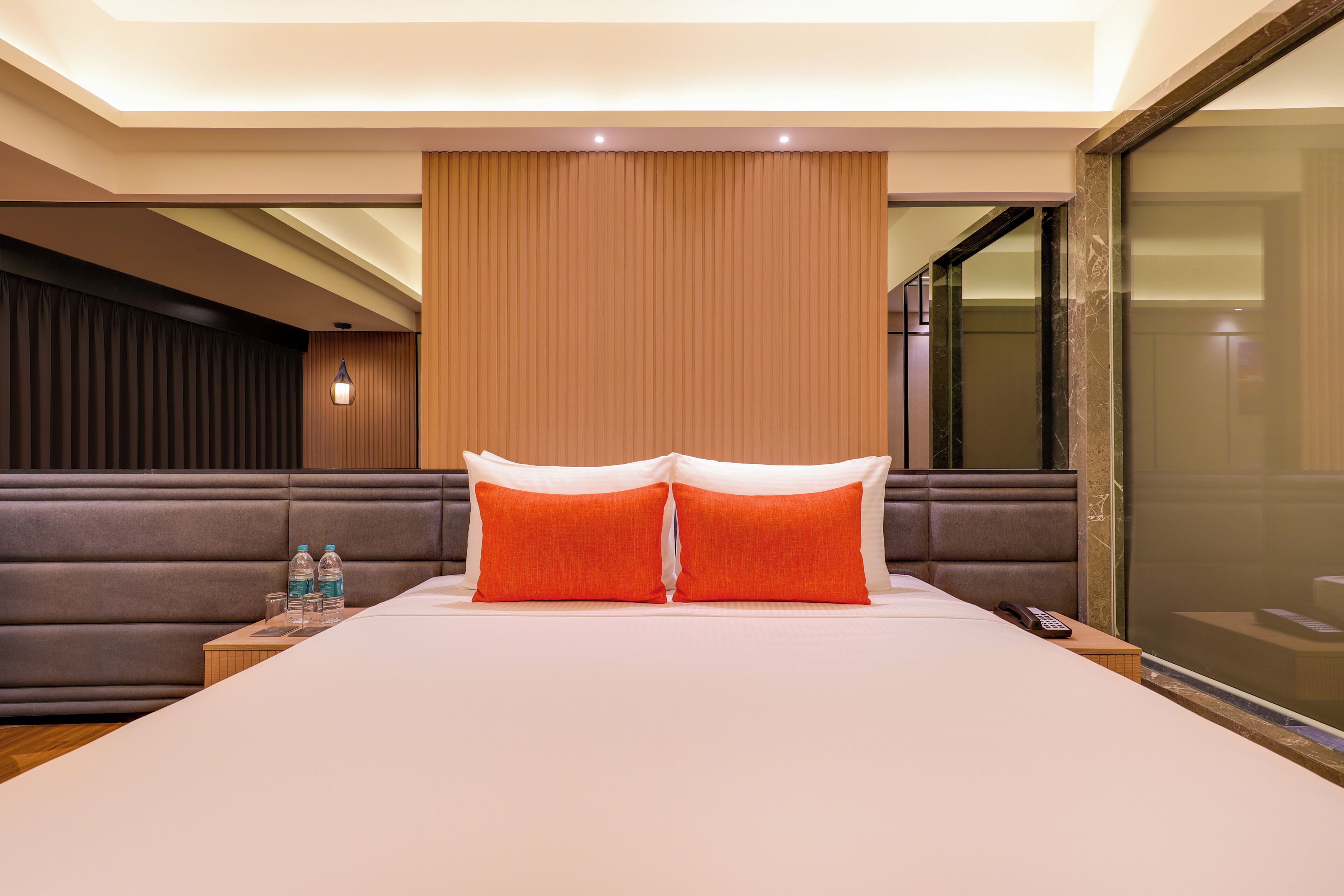 A hotel room with a large bed, two pillows, a nightstand with a phone and water bottles, a wooden headboard, and a glass partition.