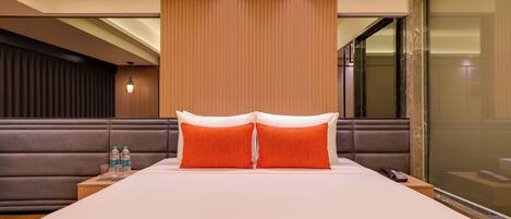 A hotel room with a large bed, two pillows, a nightstand with a phone and water bottles, a wooden headboard, and a glass partition.