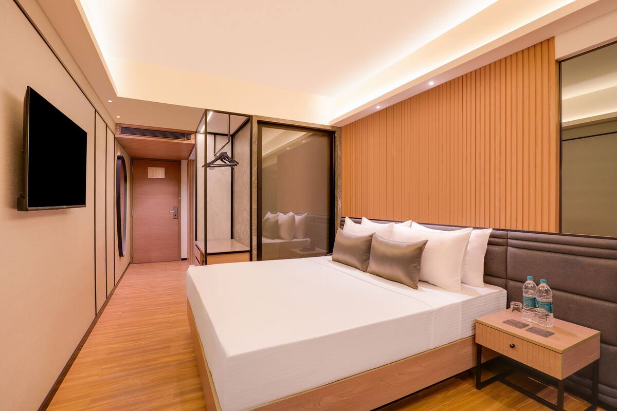 A modern hotel room with a large bed, a bedside table with a bottle and glass, and a TV mounted on the wall.