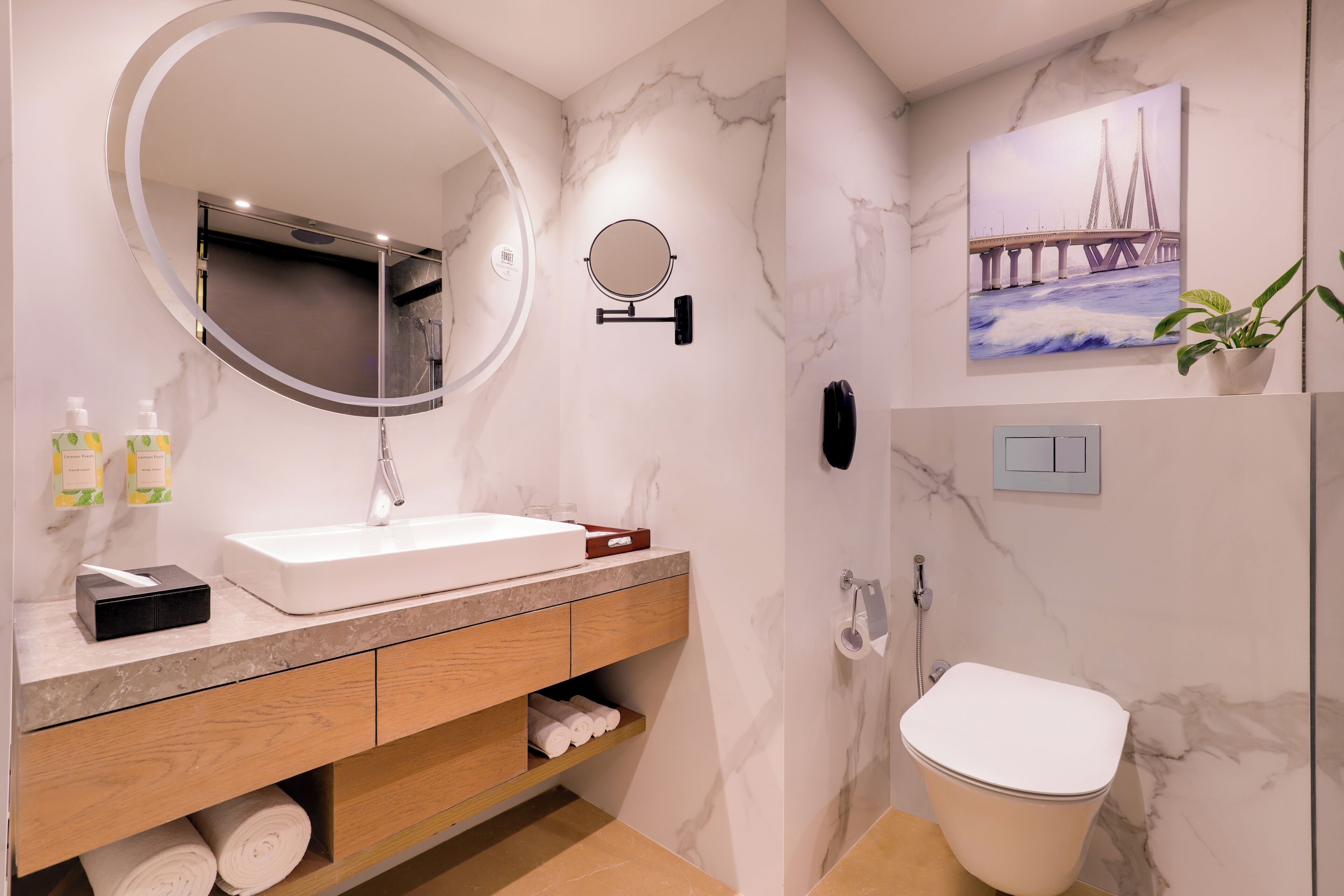 Een moderne badkamer met een marmeren wand, een grote ronde spiegel, een witte wastafel, een toilet en een douche.